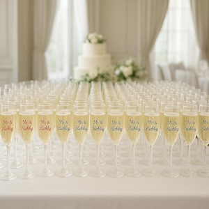May include: A table set with numerous champagne flutes filled with a golden liquid. Each glass is labeled with "Mr. & Richley" in black or red lettering, suggesting a wedding or celebratory event. A tiered cake and floral arrangements are in the background.