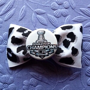 May include: A white and black leopard print bow with a button featuring the 2012 Stanley Cup Champions logo. The logo is silver and black with the text "2012 Stanley Cup Champions LA" on a white background.