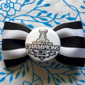 May include: Black and white striped ribbon bow with a white button featuring a silver Stanley Cup trophy and the text "2012 Stanley Cup Champions LA".