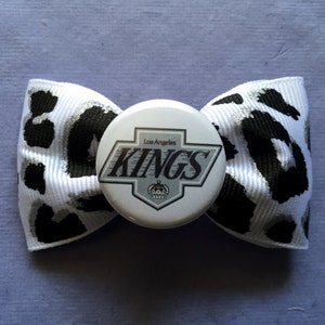 May include: A white and black leopard print bow tie with a Los Angeles Kings button in the center. The button has a silver and white logo with the words "Los Angeles Kings" and a crown.