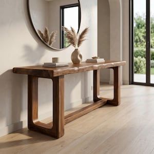 May include: A long, rectangular console table crafted from dark brown wood with a natural live edge top and two sturdy, open-frame legs connected by a horizontal base support. The table surface holds two stacks of books and a beige ceramic vase.