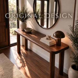 Può includere: Un lungo tavolo console in legno con bordi arrotondati e quattro gambe cilindriche. Il tavolo sostiene un vaso bianco con vegetazione, una piccola ciotola, due libri impilati e una lampada a forma di fungo. Il testo ORIGINAL DESIGN è sopra.