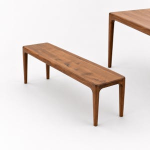Op de afbeelding: Houten bank met een rechthoekige zitting en vier gebogen poten. De bank is gemaakt van lichtbruin hout en heeft een gladde afwerking. De bank is ongeveer 152 cm lang.