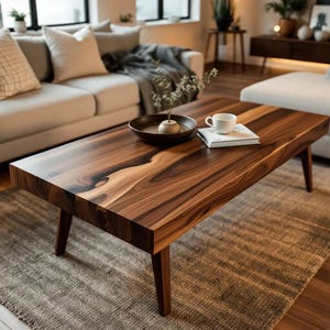 Puede incluir: Una mesa de centro rectangular de madera marrón oscuro. La superficie de la mesa muestra un patrón de vetas de madera natural y se apoya en cuatro patas cónicas. Un cuenco, un jarrón, una taza y un libro están sobre la mesa. La mesa está sobre una alfombra de color neutro.