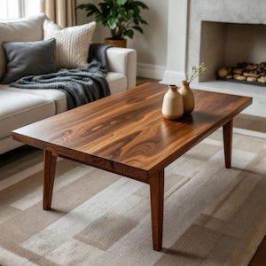 Puede incluir: Mesa de centro rectangular de madera marrón oscuro. La superficie lisa descansa sobre cuatro patas cónicas. Dos pequeños jarrones beige con flores secas están encima. La mesa está en un salón, sobre una alfombra.