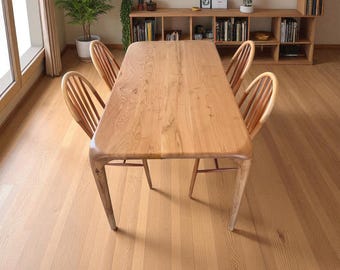 Op de afbeelding: Een lichtgekleurde houten eettafel met vier bijpassende stoelen. De tafel heeft een glad, rechthoekig oppervlak met afgeronde hoeken en wordt ondersteund door vier schuine poten. De stoelen hebben een lattenrug.