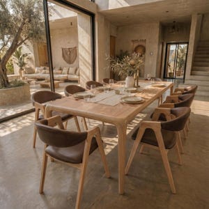 May include: A rectangular light wood dining table with rounded corners and eight matching chairs featuring light wood frames and dark brown upholstered seats. The table is set with white plates, clear glasses, and a central vase with dried foliage.