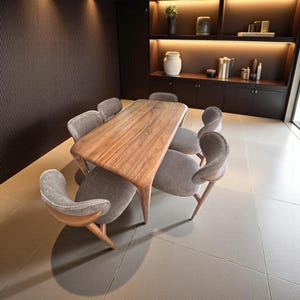 Puede incluir: Una mesa de comedor de madera con acabado natural, rodeada de seis sillas tapizadas grises. Las sillas tienen respaldos curvos y patas de madera. La habitación tiene paneles de madera oscura y una estantería empotrada con objetos decorativos.