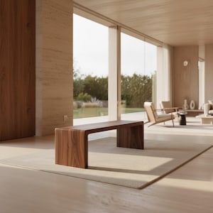 May include: A modern, solid wood bench with a curved design. The bench is a rich brown color and sits on a neutral-toned rug in a well-lit room. The room features large windows and minimalist furniture.