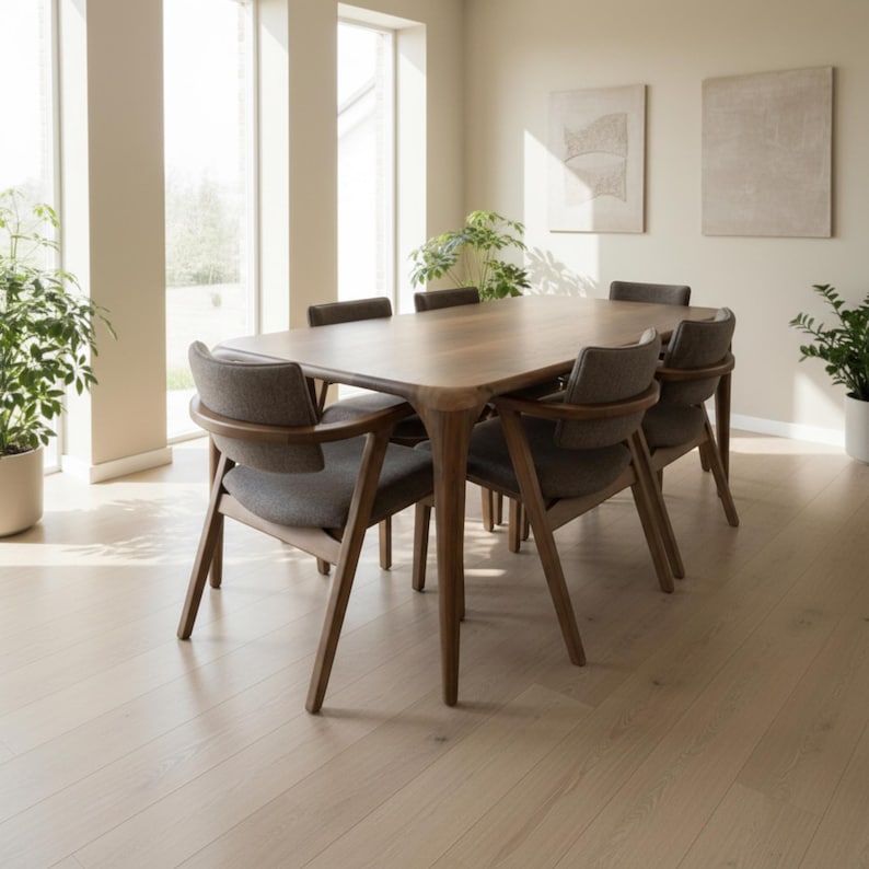 May include: A wooden dining table and six chairs. The table has a light brown surface and rounded edges. The chairs have grey upholstered seats and backs, with wooden frames. The room has light-coloured walls and flooring.