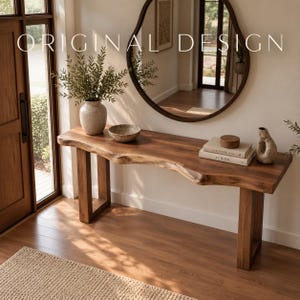 Puede incluir: Una mesa consola de madera con borde natural, acabado de veta visible y dos patas rectangulares. Sobre la mesa hay un jarrón de cerámica con follaje verde, un cuenco pequeño, dos libros apilados titulados Kinfolk y At Home, y un jarrón de cerámica texturizada.