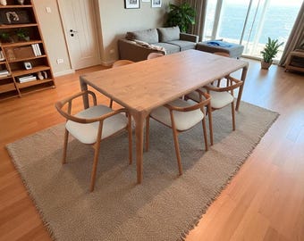 May include: A light-colored wooden dining table with six matching chairs. The chairs have light-colored seats and wooden frames. The table and chairs are on a beige rug. The room has hardwood floors and a large window.