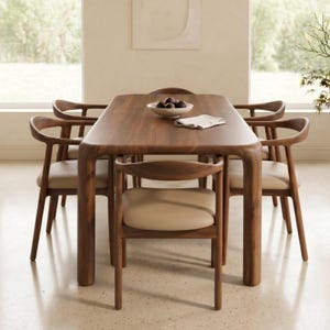 Puede incluir: Una mesa de comedor rectangular de madera con esquinas redondeadas y cuatro sillas de madera a juego. Cada silla presenta un respaldo curvo y un asiento tapizado en color beige claro. Un cuenco blanco con objetos oscuros está sobre la mesa.