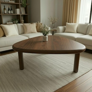 Könnte beinhalten: Ein dreieckiger Couchtisch aus dunkelbraunem Holz mit abgerundeten Kanten und drei zylindrischen Beinen. Der Tisch steht auf einem hellen Teppich in einem Wohnzimmer. Eine kleine Vase mit Blumen steht auf dem Tisch. (Ca. 100 cm)