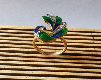 May include: A gold-toned ring featuring a peacock design. The peacock's feathers are detailed with green and blue enamel, accented with small, clear gemstones. The ring is set against a wooden background.