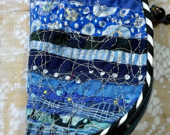 May include: A blue and black fabric eyeglass case with a zipper closure and a white and black striped trim. The case is made of patchwork fabric and has a quilted design. A pair of black eyeglasses is sticking out of the case.
