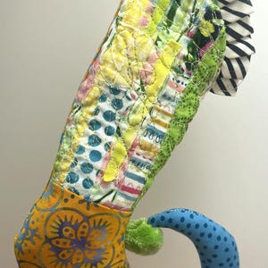 May include: A whimsical, handmade fabric seahorse with a patchwork design. The seahorse features a yellow and blue patterned body, a green pom-pom, and a striped fin. The colorful design is made of various fabrics.