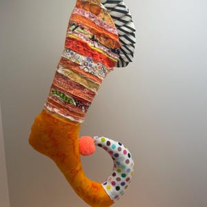 May include: A whimsical, handmade Christmas stocking with a patchwork design in shades of orange, red, and yellow. The foot is bright orange, and the curled toe features a polka dot pattern. A small orange pom-pom is attached.