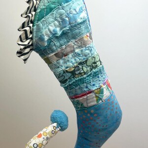 May include: A whimsical, handmade Christmas stocking with a unique, curved design. The stocking features a patchwork of blue fabrics, a striped ruffle, and a patterned, curled toe. A small, blue pom-pom adorns the toe.