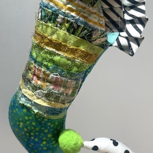 May include: A whimsical, handmade fabric seahorse. The body is a patchwork of colorful fabrics, with a green and blue spotted tail. The seahorse has a green pom-pom and a curved tail with black polka dots.