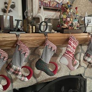 May include: Five red and white striped stockings with elf shoe bottoms are hung on a wooden mantel. The stockings are made of a patchwork fabric with plaid and gingham patterns. The stockings are hung with hooks that are shaped like the letters "J", "O", and a fleur-de-lis.