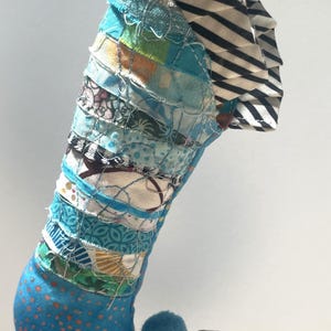 May include: A handmade Christmas stocking with a whimsical design. The body is a patchwork of blue, green, and patterned fabrics, with a blue pom-pom and a striped cuff. The toe is a curved shape with colorful circles.