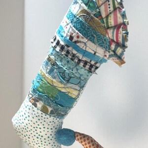 May include: A whimsical, handmade fabric ornament with a unique shape. The ornament features a combination of patterned fabrics in blue, white, and orange, with a blue pom-pom and a dotted pattern. The top is pleated.