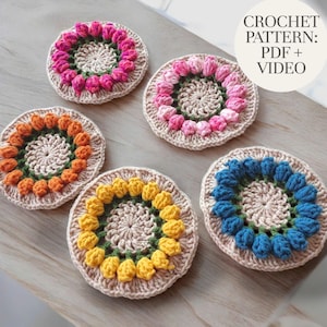May include: Five handmade crochet coasters in various colours: pink, orange, yellow, and blue. Each coaster features a floral design with a central circle and a ring of flower-shaped embellishments. The text "CROCHET PATTERN: PDF + VIDEO" is visible.