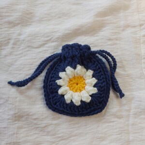 May include: A small, square, dark blue crochet drawstring pouch featuring a central daisy motif. The flower has a bright yellow center surrounded by white petals, all crafted from yarn. The pouch includes two dark blue braided drawstrings.