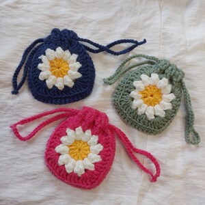 May include: Three small crochet drawstring pouches in dark blue, sage green, and bright pink. Each pouch features a central daisy motif with a yellow center and white petals, finished with matching crocheted drawstring cords.