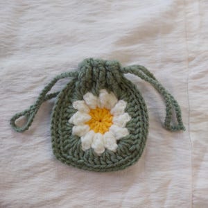 May include: A small, round, sage green crochet drawstring pouch featuring a central daisy motif with white petals and a bright yellow center. The pouch is constructed with thick yarn and includes two matching green crocheted drawstrings.