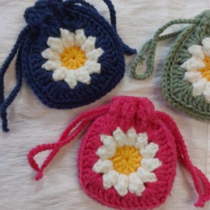 May include: Three small, round, crocheted drawstring pouches in dark blue, sage green, and bright pink. Each pouch features a central daisy motif with white petals and a yellow center, finished with matching crocheted drawstrings.