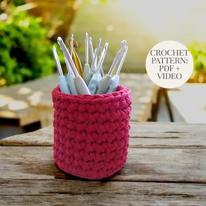 May include: A magenta-coloured crochet hook holder, handmade from thick yarn, filled with various crochet hooks. The cylindrical holder has a textured appearance. A white circle in the background reads "CROCHET PATTERN: PDF + VIDEO".