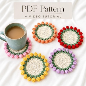 May include: A light blue ceramic mug sits on a round, cream-coloured crochet coaster with a green border and pink petal-like edges. Four additional coasters in orange, red, yellow, and purple are arranged nearby. Text reads: PDF Pattern + VIDEO TUTORIAL.