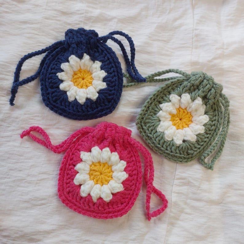 May include: Three small drawstring pouches made of crocheted yarn. Each pouch features a central daisy motif with white petals and a yellow center. The pouches are colored in dark blue, sage green, and bright pink, each with matching drawstrings.