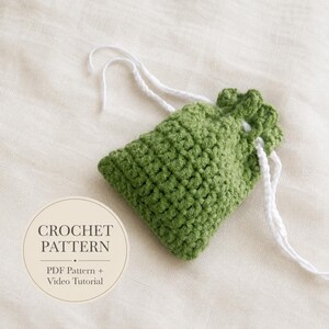 Puede incluir: Una pequeña bolsa de ganchillo verde con cordón, patrón de puntos texturizados y lazos de hilo blanco. Una superposición circular a la izquierda muestra el texto CROCHET PATTERN seguido de PDF Pattern + Video Tutorial en letra oscura.