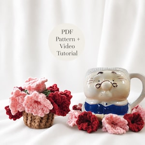 Puede incluir: Un arreglo floral de ganchillo en una maceta marrón y una taza de cerámica con forma de persona con gafas, rodeado de flores de ganchillo rosas y rojo oscuro. El texto PDF Pattern + Video Tutorial es visible en el centro de la imagen.