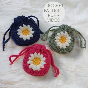 May include: Three small crochet drawstring pouches in navy blue, sage green, and bright pink. Each pouch features a central white and yellow daisy motif. Text in the image reads: CROCHET PATTERN: PDF + VIDEO.