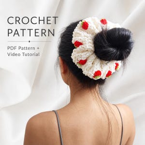Może przedstawiać: Biała szydełkowa gumka do włosów z małymi czerwonymi motywami truskawek z zielonymi listkami. Tekst CROCHET PATTERN PDF Pattern + Video Tutorial jest widoczny po lewej stronie. Gumka jest założona na ciemny kok, ukazując fakturę włóczki.
