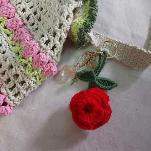 Peut inclure: Sac crocheté beige avec des motifs floraux roses et verts. Un porte-clés en forme de rose rouge crochetée avec des feuilles vertes et un anneau doré est attaché. Le sac présente un motif floral.