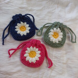 May include: Three small crochet drawstring pouches in navy blue, sage green, and bright pink. Each pouch features a central daisy motif with white petals and a yellow center, constructed with a thick yarn texture and matching drawstring cords.
