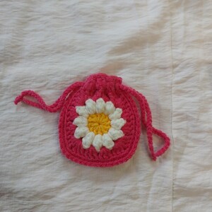 May include: A small, round, crocheted drawstring pouch in bright pink yarn. The center of the pouch features a prominent, raised daisy motif with a vibrant yellow center surrounded by white petals, all crafted from yarn.