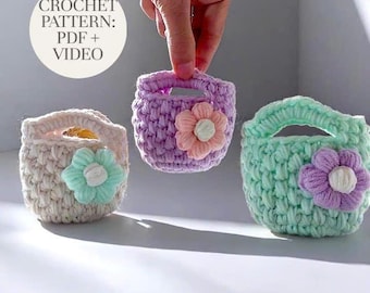 Könnte beinhalten: Drei kleine gehäkelte Körbchen mit Henkeln, jeweils mit einer Garnblumenapplikation. Die Körbchen sind beige, lila und mintgrün. Der Text auf dem Bild lautet: CROCHET PATTERN: PDF + VIDEO. Diese Artikel sind handgefertigt aus Garn.