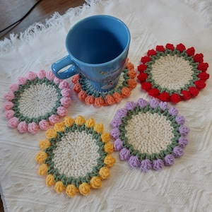 May include: A blue ceramic mug rests on a crocheted coaster. The coasters are circular, with a cream-colored center and colorful, flower-shaped edges in pink, orange, red, yellow, and purple. The coasters are handmade and sit on a white, textured surface.
