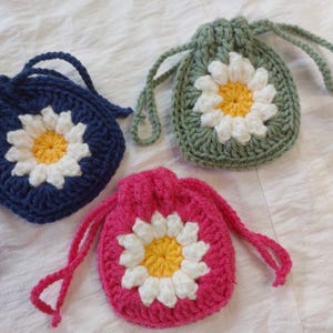 May include: Three small, round, crocheted drawstring pouches. One is dark blue, one is sage green, and one is bright pink. Each pouch features a central daisy motif with white petals and a yellow center, crafted from yarn.