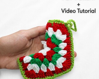 May include: A crochet stocking ornament featuring a granny square pattern in red, white, and green yarn with a small loop at the top. The text PDF Pattern + Video Tutorial is printed in black above the crocheted item on a white background.