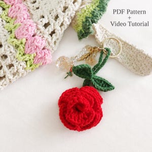 May include: A crochet keyring featuring a vibrant red rose with green leaves attached to a gold-tone metal ring. The accessory includes clear beads and a small rose charm. Text in the image reads: PDF Pattern + Video Tutorial.