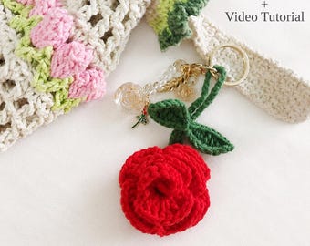 Peut inclure: Un porte-clés en crochet représentant une rose rouge vive avec des feuilles vertes, fixé à un anneau en métal doré. L'accessoire comprend des perles transparentes et une petite breloque rose. Texte : PDF Pattern + Video Tutorial.