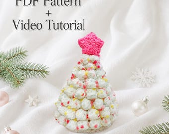 Könnte beinhalten: Ein gehäkelter Weihnachtsbaumschmuck aus weißem Garn mit bunten Sprenkeln und einem leuchtend rosa gehäkelten Stern als Spitze. Der Text PDF Pattern + Video Tutorial steht über dem Baum. Der Baum ist von Tannenzweigen und Kugeln umgeben.