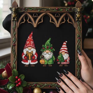 Puede incluir: Obra de arte de punto de cruz con tres gnomos navideños en un marco de madera decorativo. Los gnomos llevan gorros rojos y verdes y sostienen regalos y linternas. El marco tiene un diseño de arco gótico con vides y gárgolas decorativas.